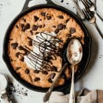 Overhead of a giant chocolate chunk skillet cookie topped with vanilla ice cream and chocolate sauce. Styled on a white background