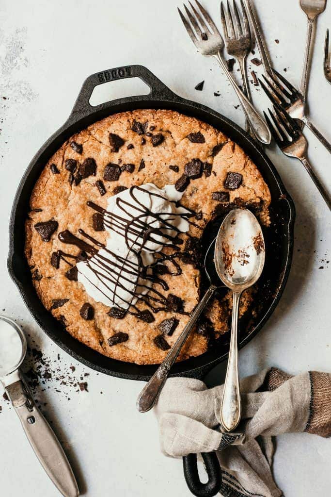 Overhead of a giant chocolate chunk skillet cookie topped with vanilla ice cream and chocolate sauce. Styled on a white background