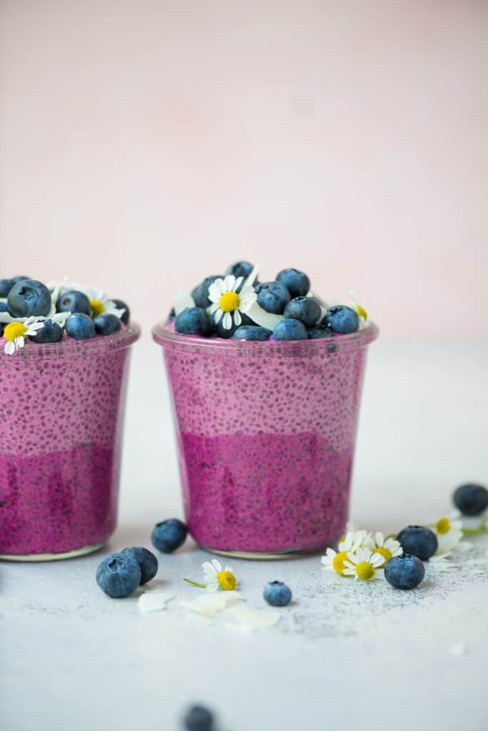 Side view of ombré pink pitaya chia pudding topped with fresh blueberries, coconut and white and yellow flowers