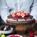 Berry vegan cheesecake with chocolate ganache and fresh fruit on a wood cake stand with person in background picking it up