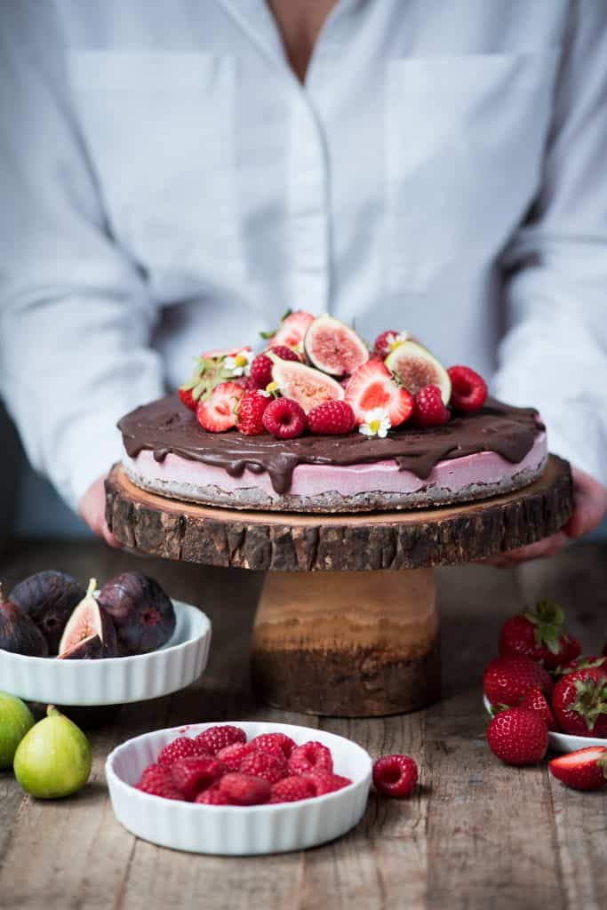Berry vegan cheesecake with chocolate ganache and fresh fruit on a wood cake stand with person in background picking it up