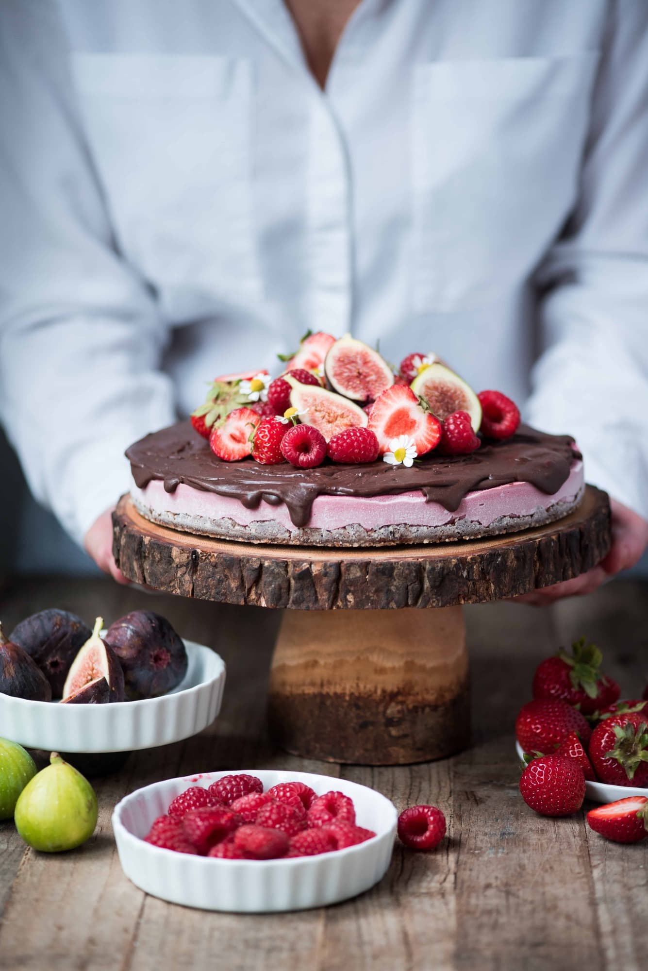 Berry vegan cheesecake with chocolate ganache and fresh fruit on a wood cake stand with person in background picking it up