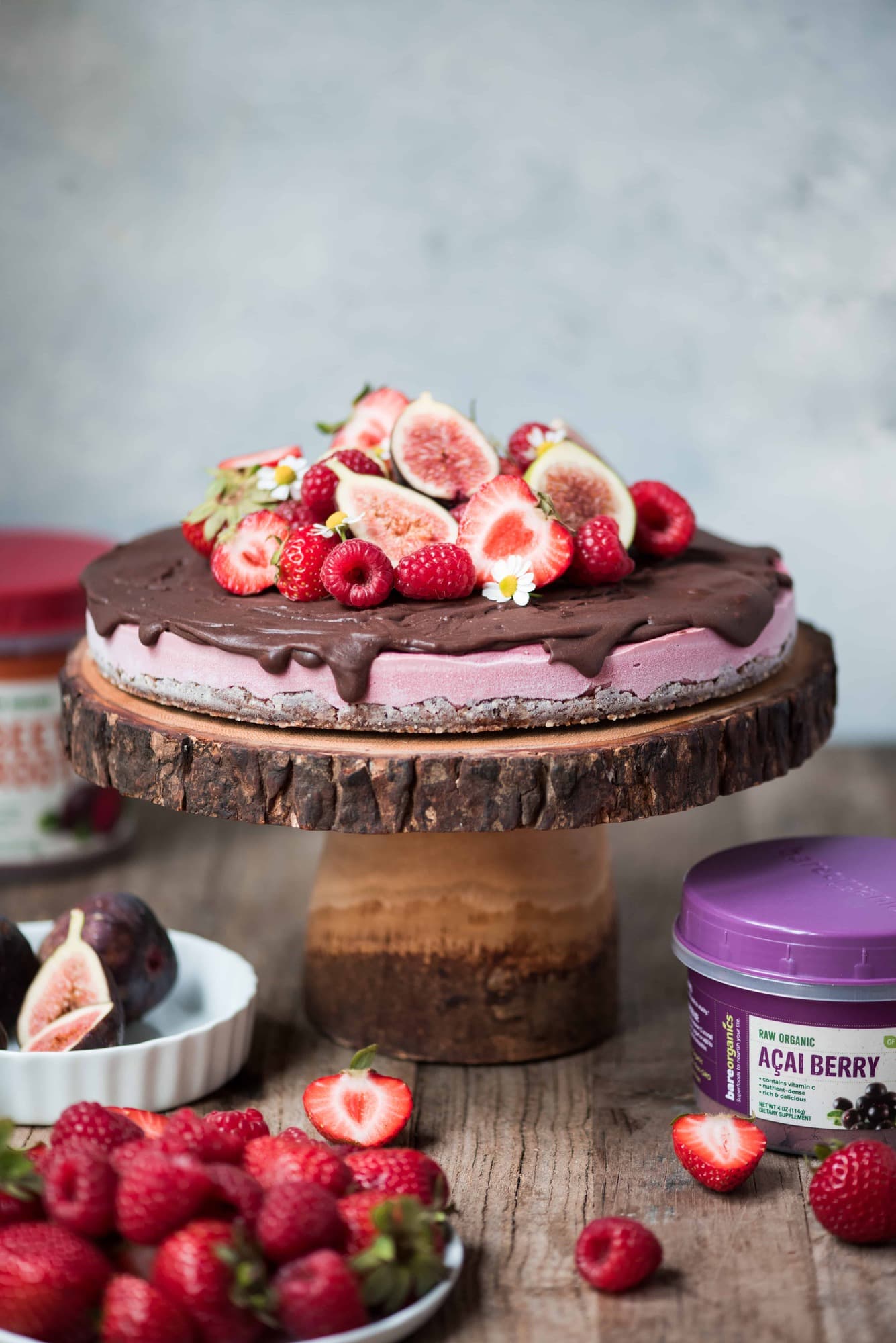 Berry vegan cheesecake with chocolate ganache and fresh fruit on a wood cake stand surrounded by bowls of fresh berries
