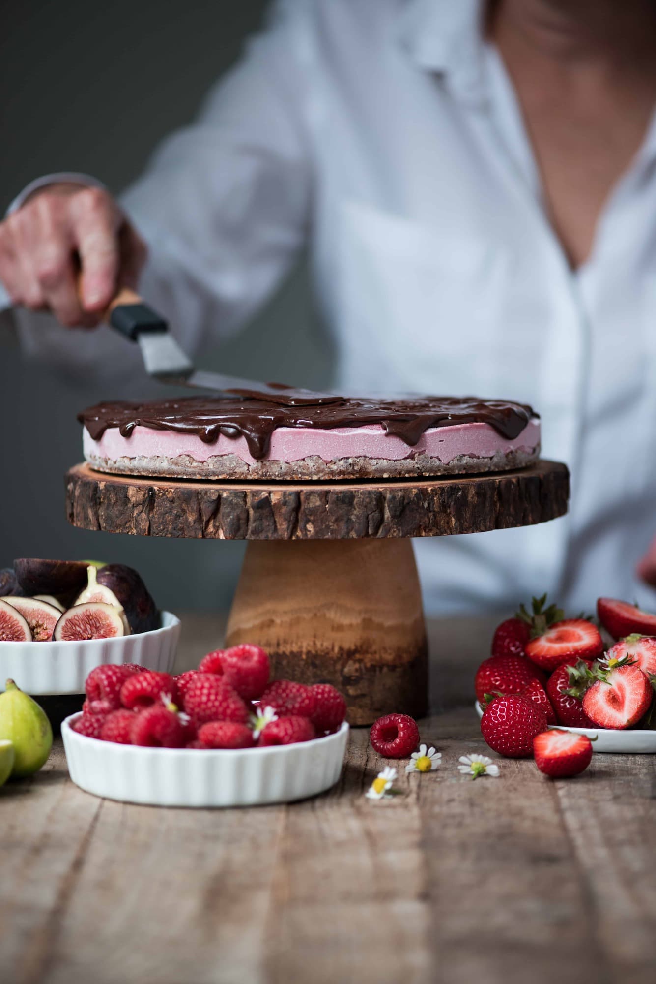 Spreading chocolate ganache on vegan berry cheesecake on wooden cake stand surrounded by bowls of fresh fruit