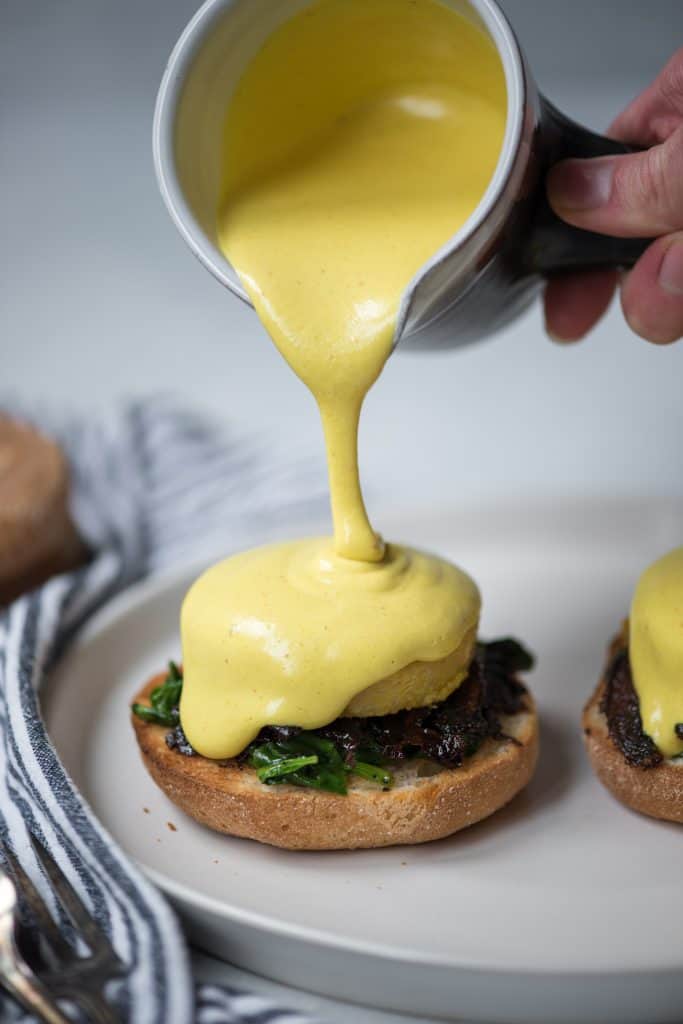 Pouring vegan hollandaise onto polenta eggs benedict on a white plate