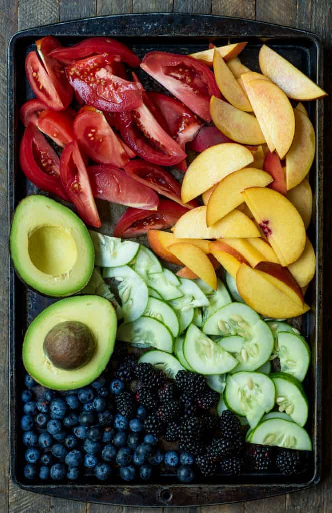 Sheet pan filled with fresh sliced tomatoes, sliced peaches, an avocado, cucumber and blueberries