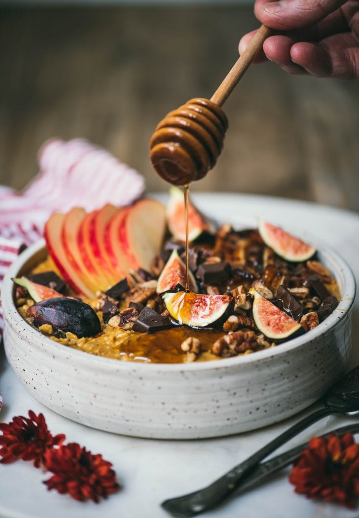 Side view of drizzling maple syrup onto a bowl of pumpkin peanut butter oatmeal topped with caramelized banana, fresh figs, apple, pecans and dark chocolate