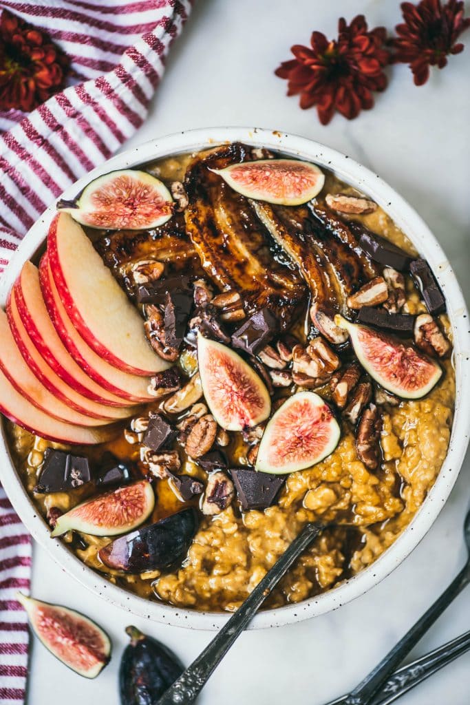 Overhead of a white bowl with pumpkin peanut butter oatmeal topped with caramelized banana, fresh figs, apple, pecans and dark chocolate