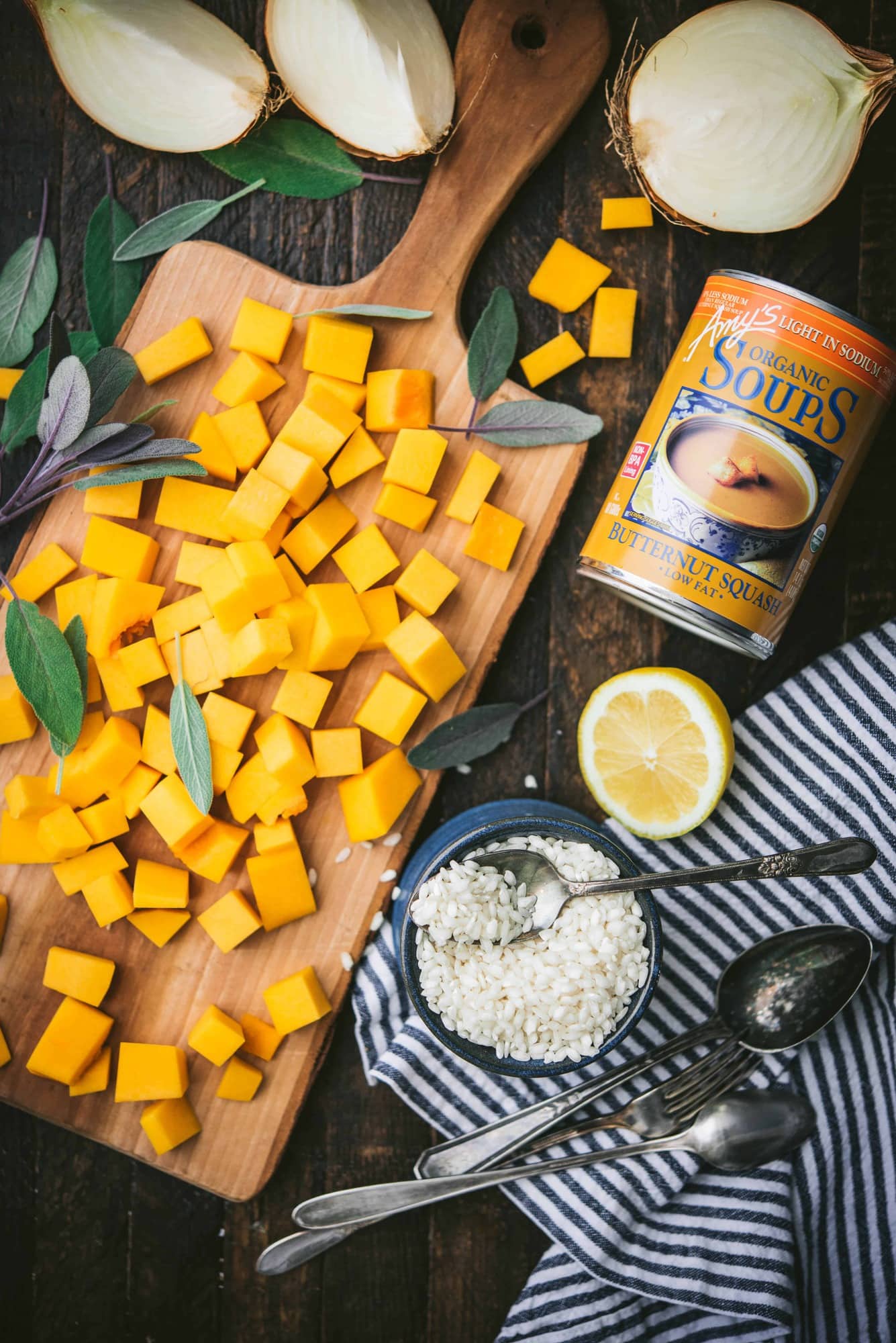 Overhead of wood cutting board with diced butternut squash, next to a bowl of arborio rice, fresh lemon and a can of Amy's butternut squash soup