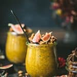 Side view of pumpkin chia pudding in a small glass jar topped with fresh figs and granola. Styled on a dark, rustic background with fall flowers in background
