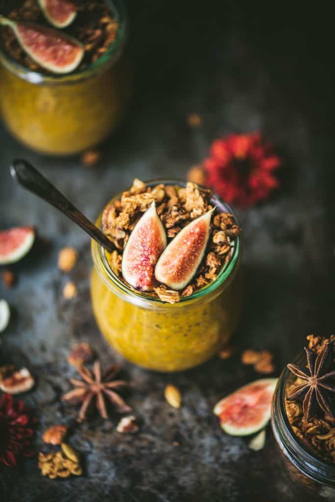 High side view of pumpkin chia pudding in a small glass jar topped with fresh figs and granola. Styled on a dark, rustic background