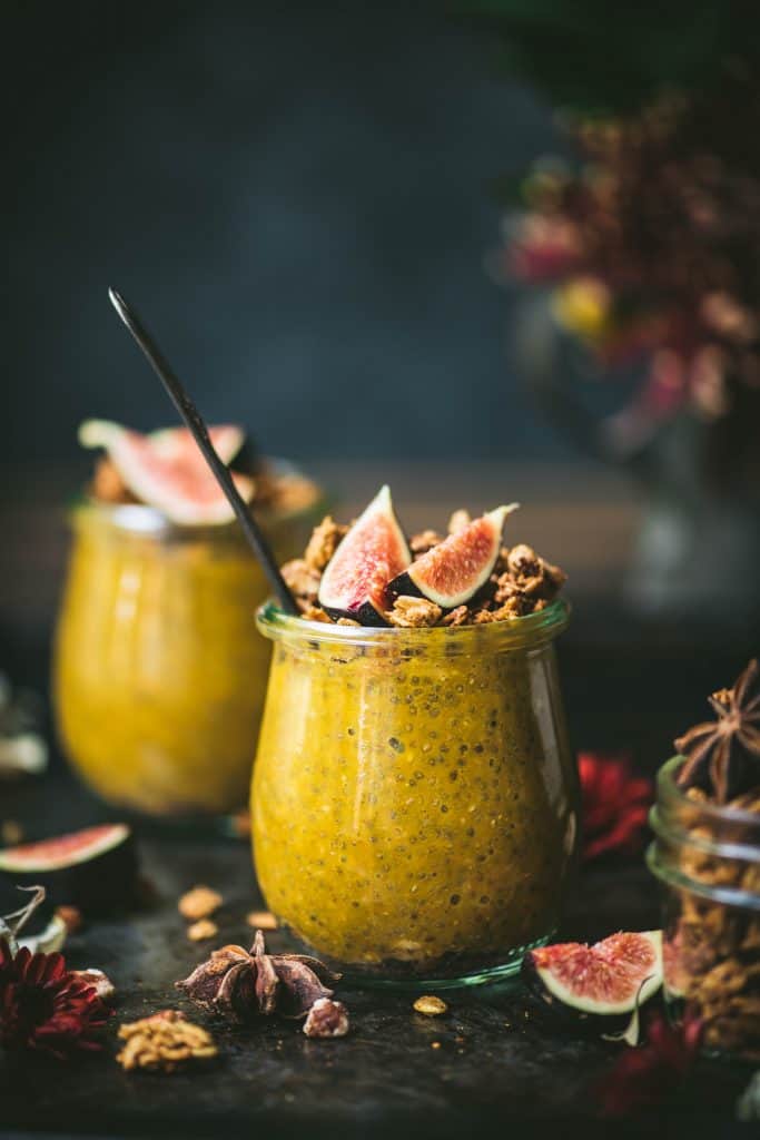 Side view of pumpkin chia pudding in a small glass jar topped with fresh figs and granola. Styled on a dark, rustic background with fall flowers in background