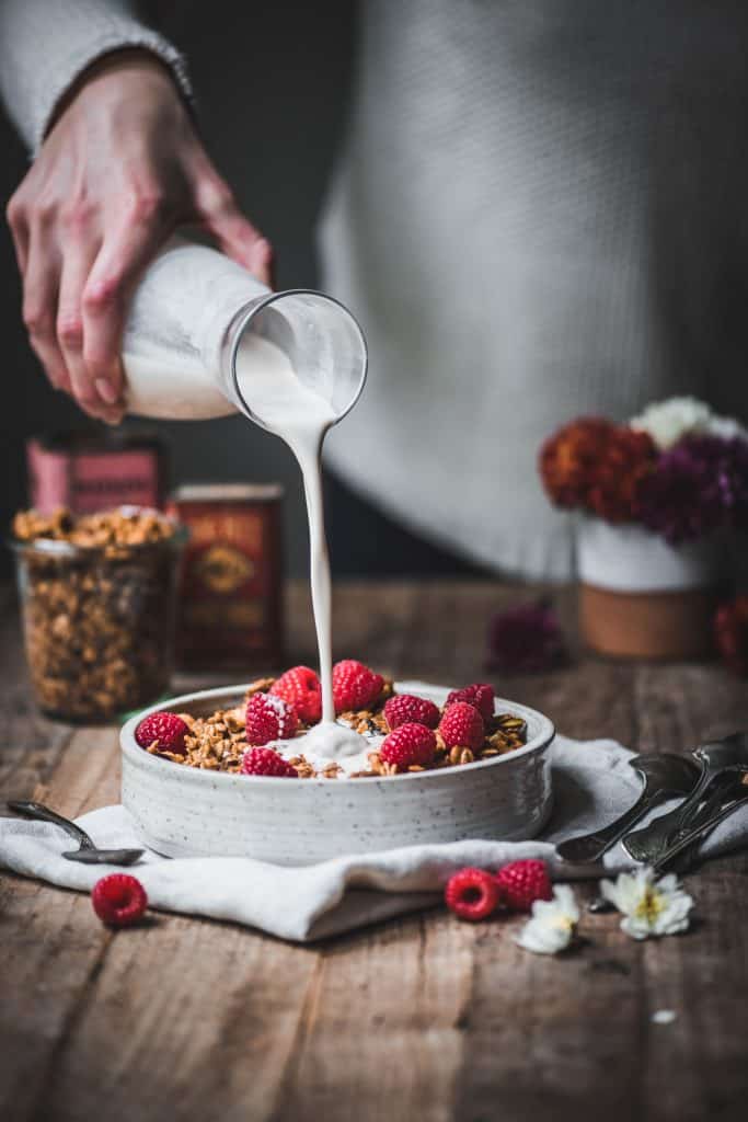Pouring milk over pumpkin spice granola with fresh raspberries on a rustic wood table