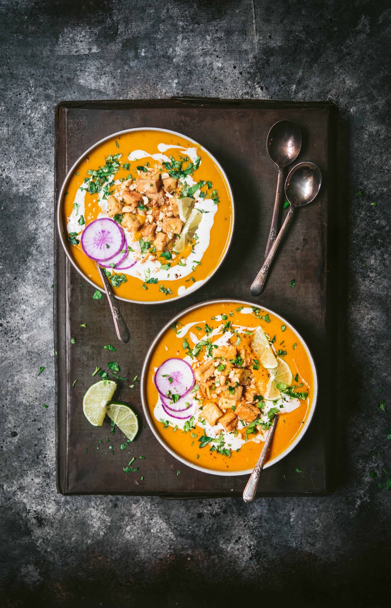 Overhead of two bowls of thai red curry pumpkin soup in black bowl garnished with crispy tofu, lime, fresh herbs and purple daikon radish