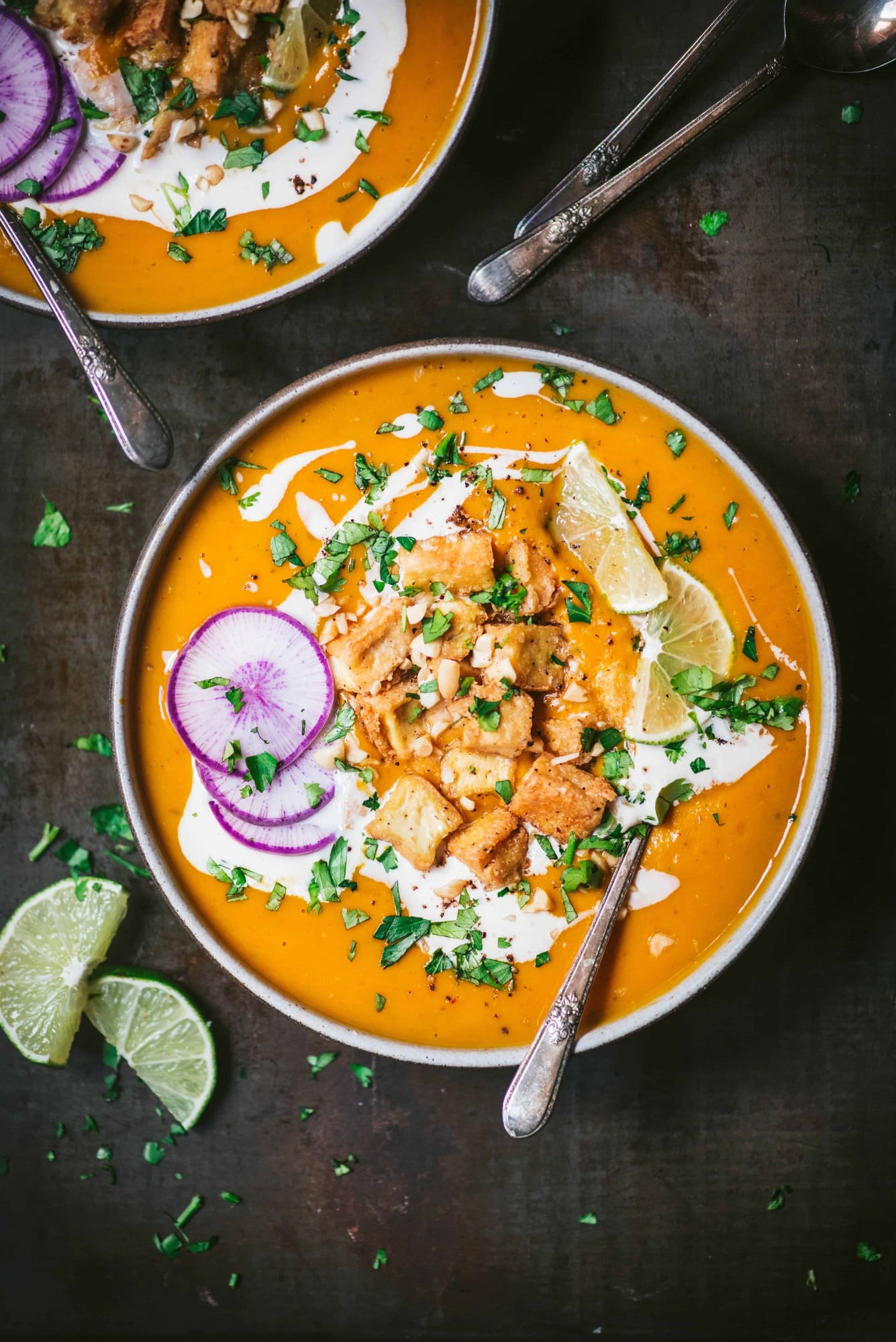 Overhead of thai red curry pumpkin soup in black bowl garnished with crispy tofu, lime, fresh herbs and purple daikon radish