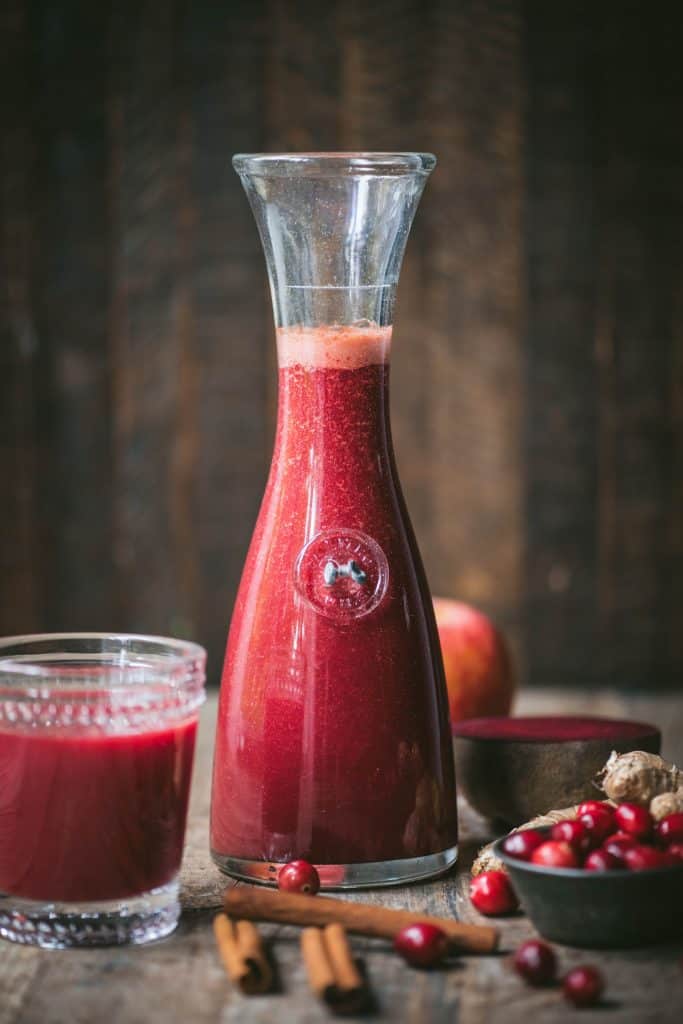 Fresh cranberry juice in a glass carafe with fresh cranberries and cinnamon sticks on rustic wooden table.