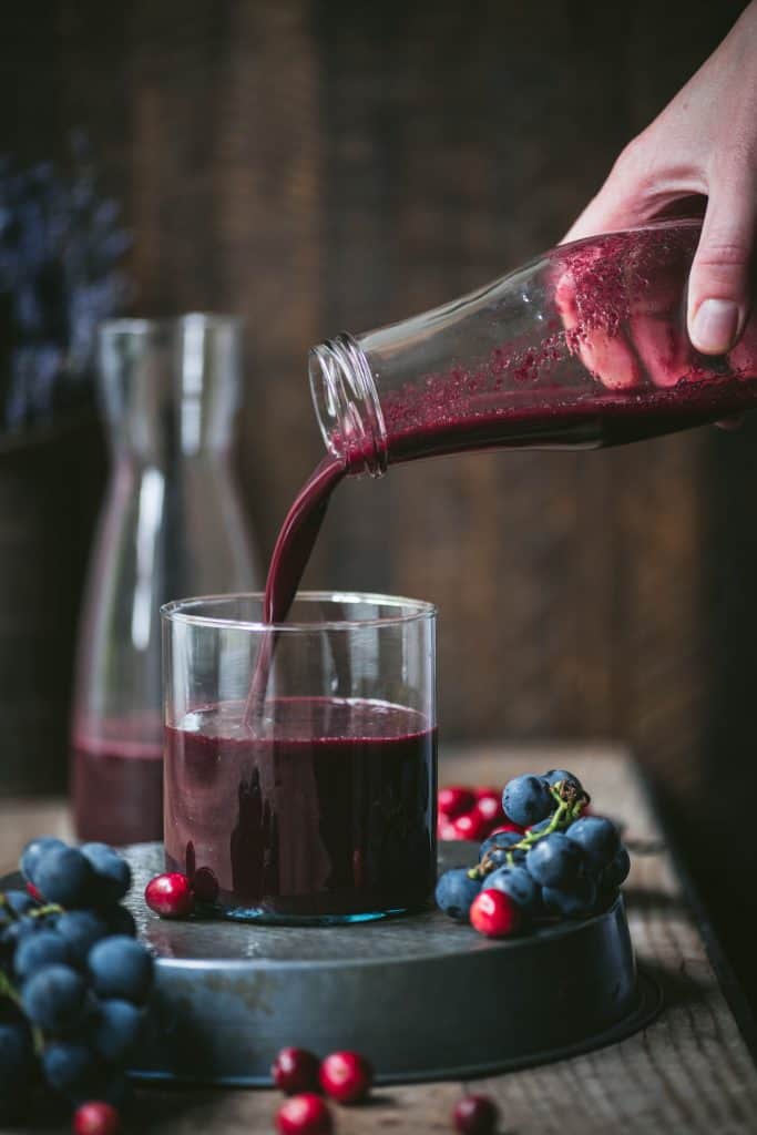 Fresh homemade berry juice being poured into a glass from a jar with red and blue berries around.