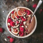 Overhead of chocolate smoothie bowl topped with fresh raspberries, figs and pomegranate in a white bowl with an antique spoon