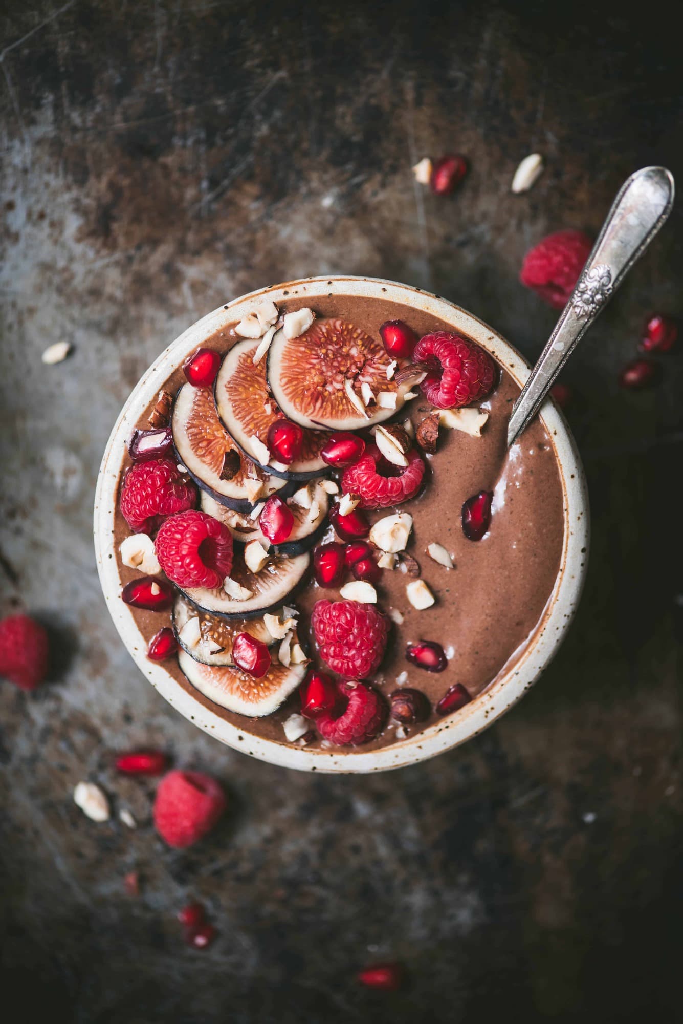Overhead of chocolate smoothie bowl topped with fresh raspberries, figs and pomegranate in a white bowl with an antique spoon