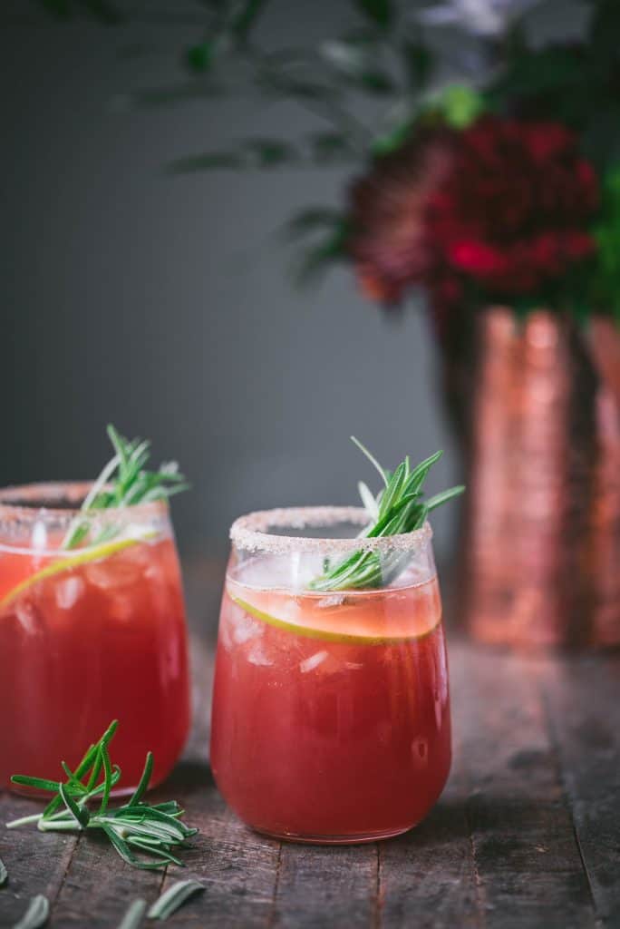 Side view of apple cider cranberry margarita with rosemary and apple garnish on rustic wood background with flowers in copper vase