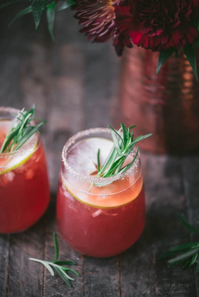 45 degree angle of apple cider cranberry margarita with rosemary and apple garnish on rustic wood background