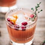 Close up of cranberry orange cocktails garnished with fresh thyme on a wood table with window in background