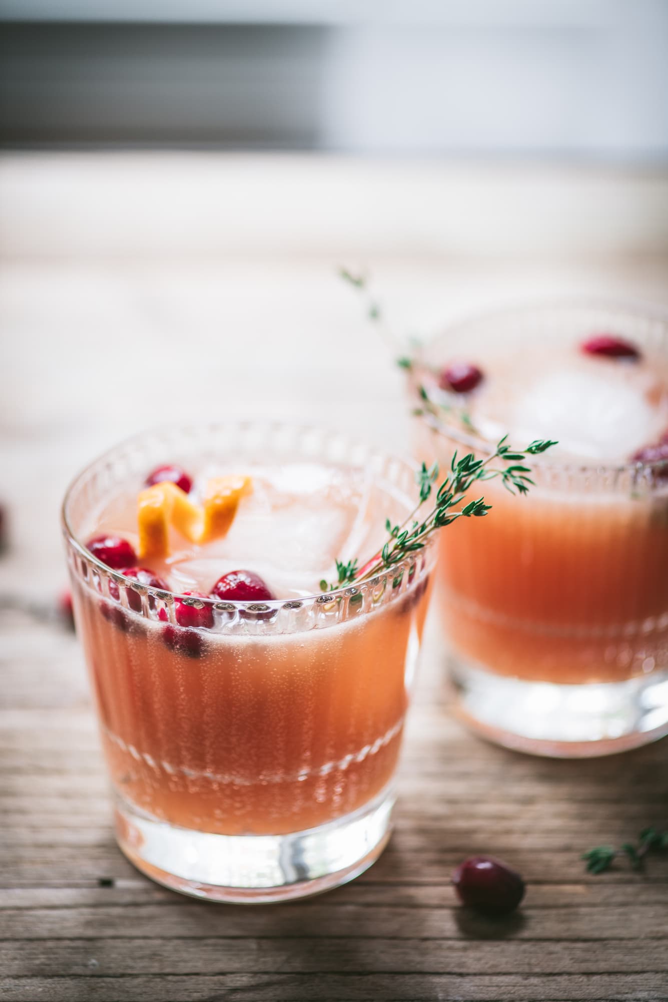 Two cranberry orange cocktails garnished with fresh thyme on a wood table with window in background