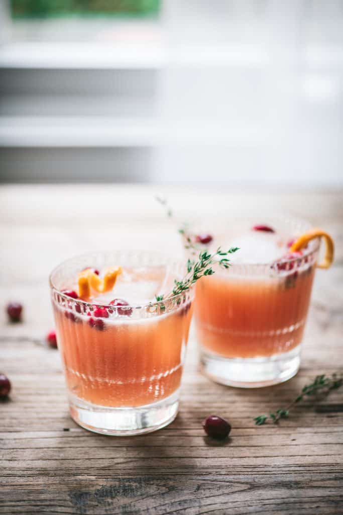 Two cranberry orange cocktails garnished with fresh thyme on a wood table with window in background