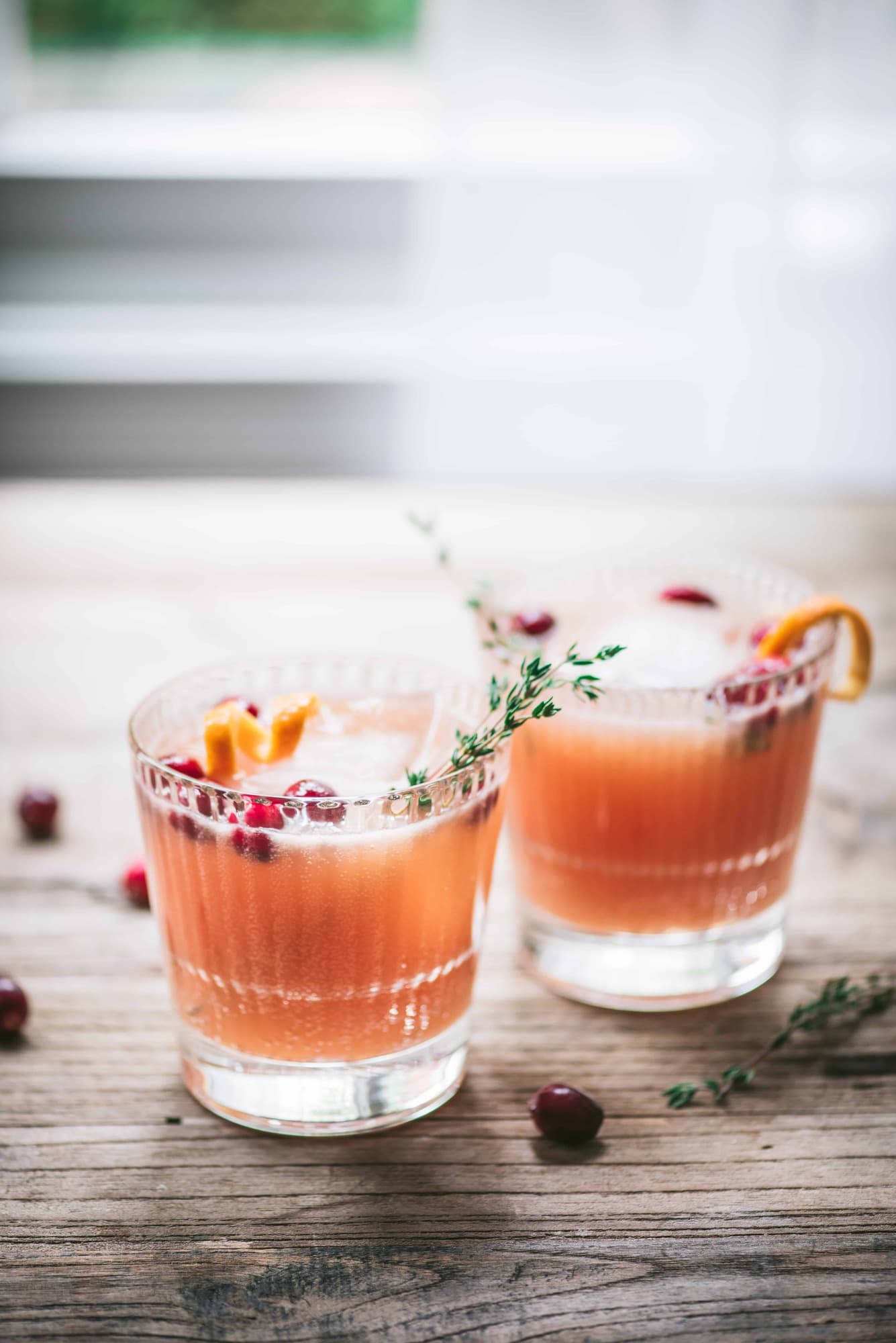 Two cranberry orange cocktails garnished with fresh thyme on a wood table with window in background