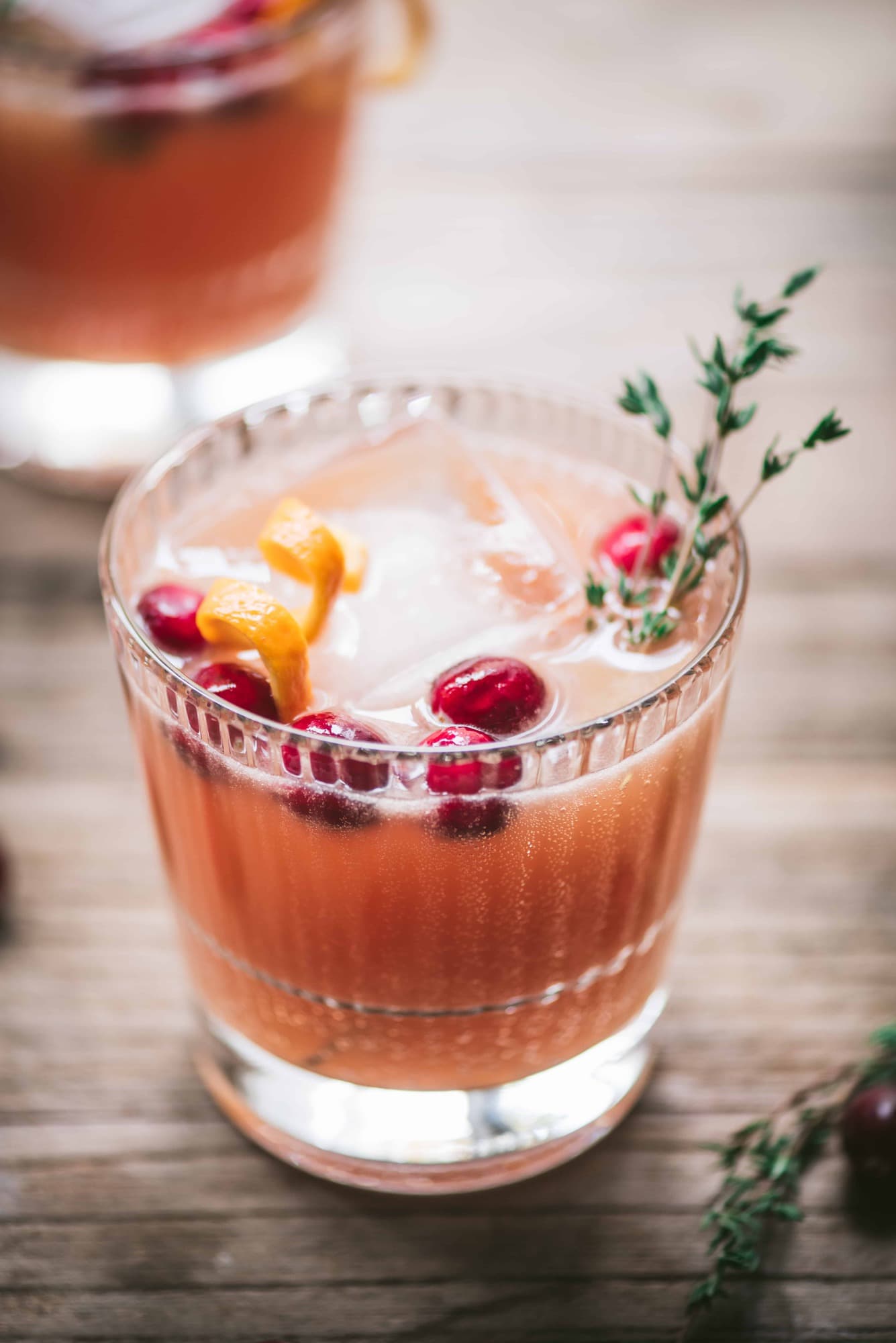 Close up of cranberry orange cocktails garnished with fresh thyme on a wood table with window in background