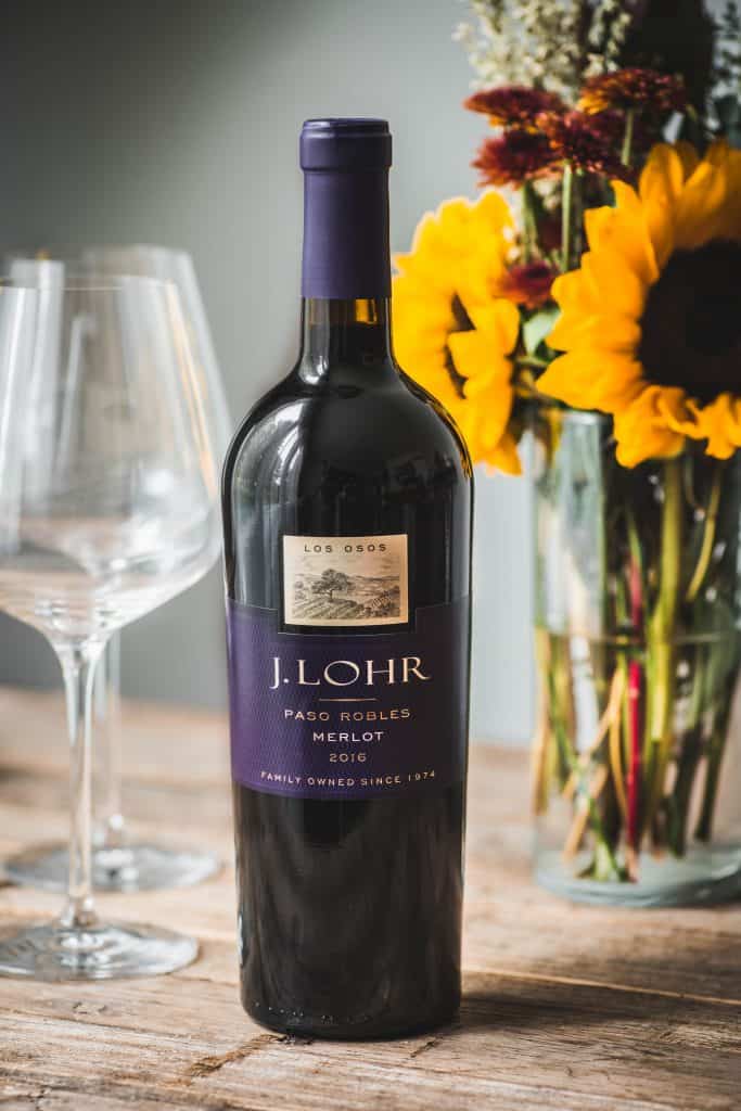 Bottle of J. Lohr Los Osos Merlot on a wooden table with wine glasses and flowers in background