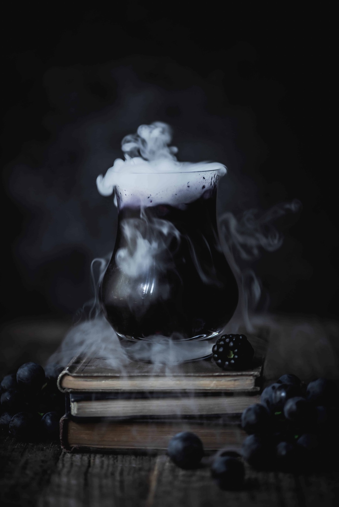 Black and purple halloween cocktail on a stack of old books with dry ice coming out of top