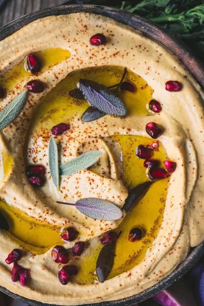 Close up of creamy pumpkin sage hummus garnished with olive oil, pomegranate seeds and fresh sage