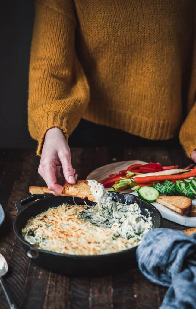 Side view of person in sweater dipping bread into vegan spinach artichoke dip in cast iron skillet