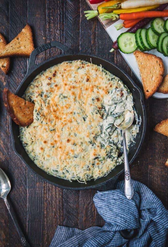 Overhead of vegan spinach artichoke dip in a cast iron skillet with toast and fresh vegetables