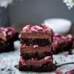 Side view of three stacked chocolate ganache brownies with rose petals on white marble