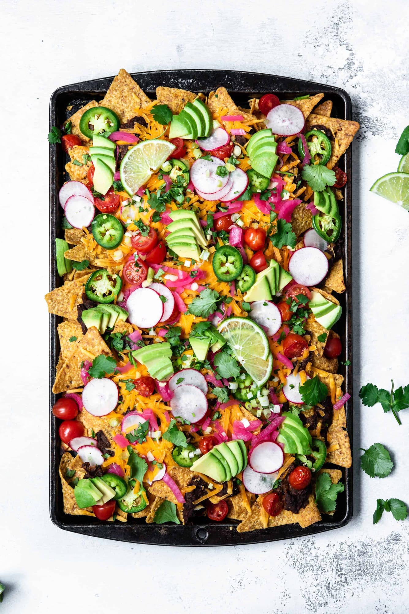 Overhead view of loaded black bean nachos on a sheet pan with avocado, cheese, pickled onion, tomatoes and jalapeño