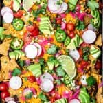 Close up Overhead view of loaded black bean nachos on a sheet pan with avocado, cheese, pickled onion, tomatoes and jalapeño
