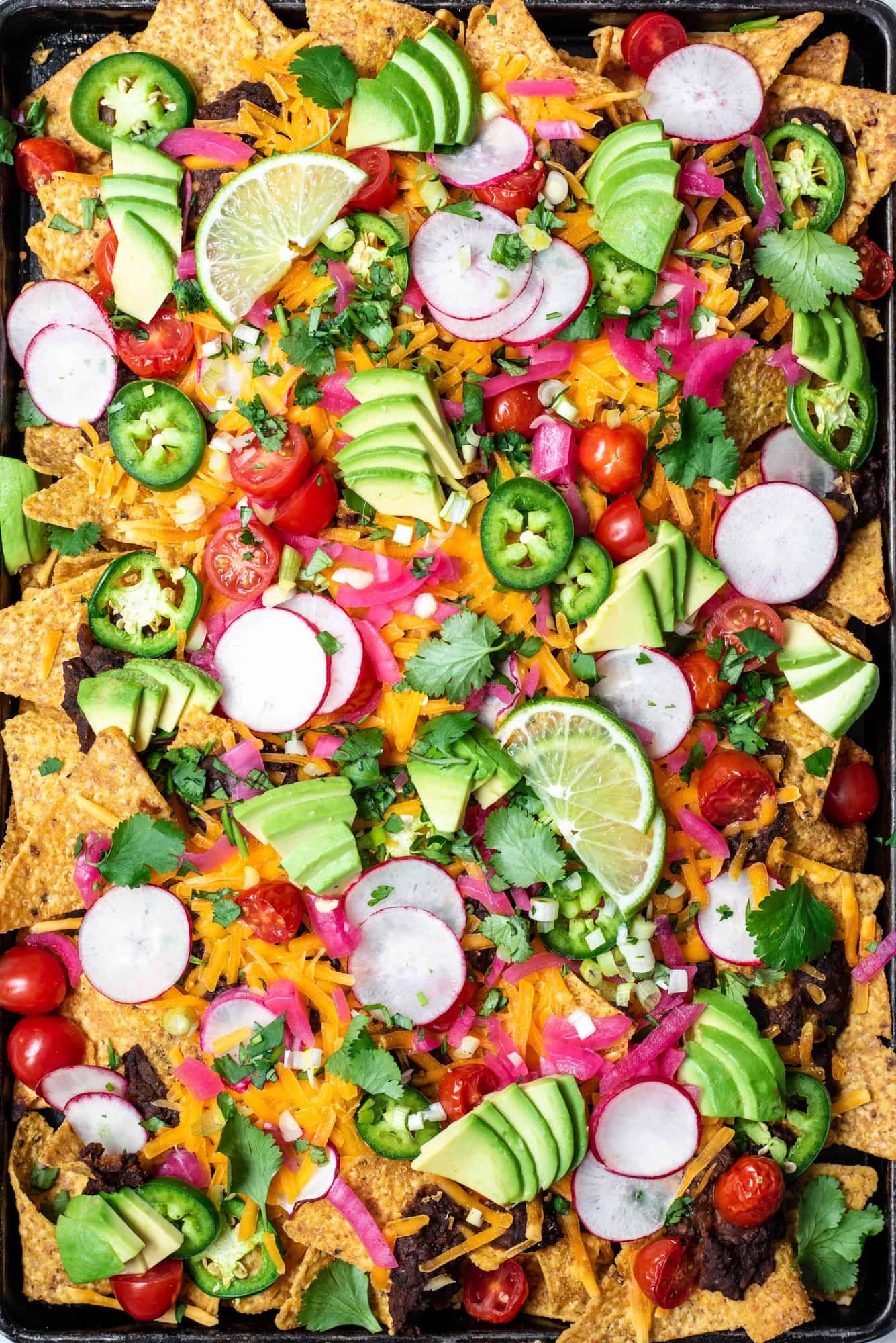 Close up Overhead view of loaded black bean nachos on a sheet pan with avocado, cheese, pickled onion, tomatoes and jalapeño