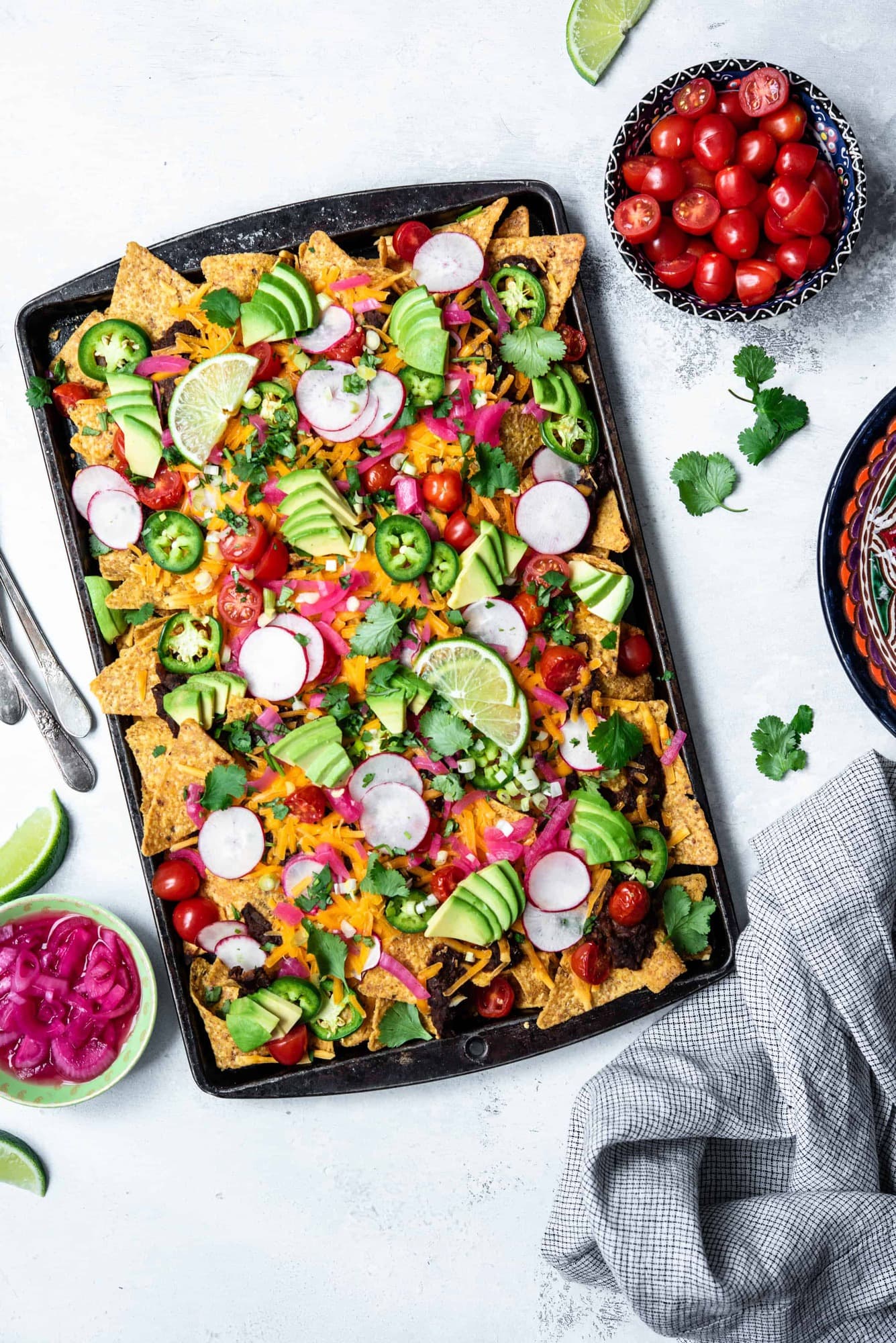 Overhead view of loaded black bean nachos on a sheet pan with avocado, cheese, pickled onion, tomatoes and jalapeño