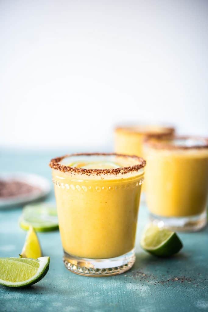Side view of mango margarita with chili salt rim and limes on teal surface