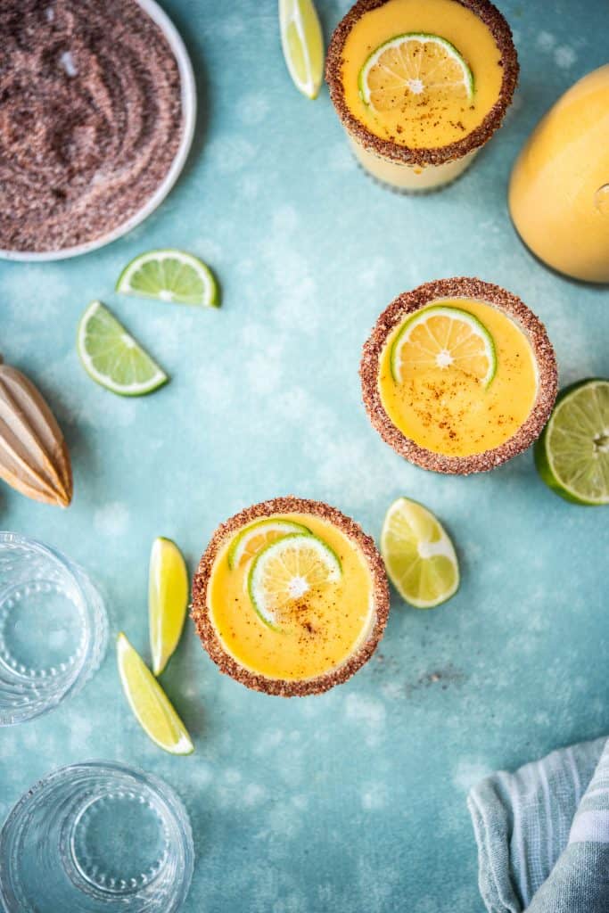 Overhead view of mango chili margaritas with limes on teal background