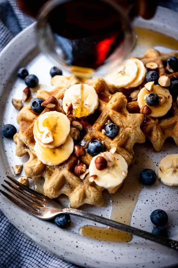 Close up view of pouring maple syrup on waffles topped with bananas and blueberries