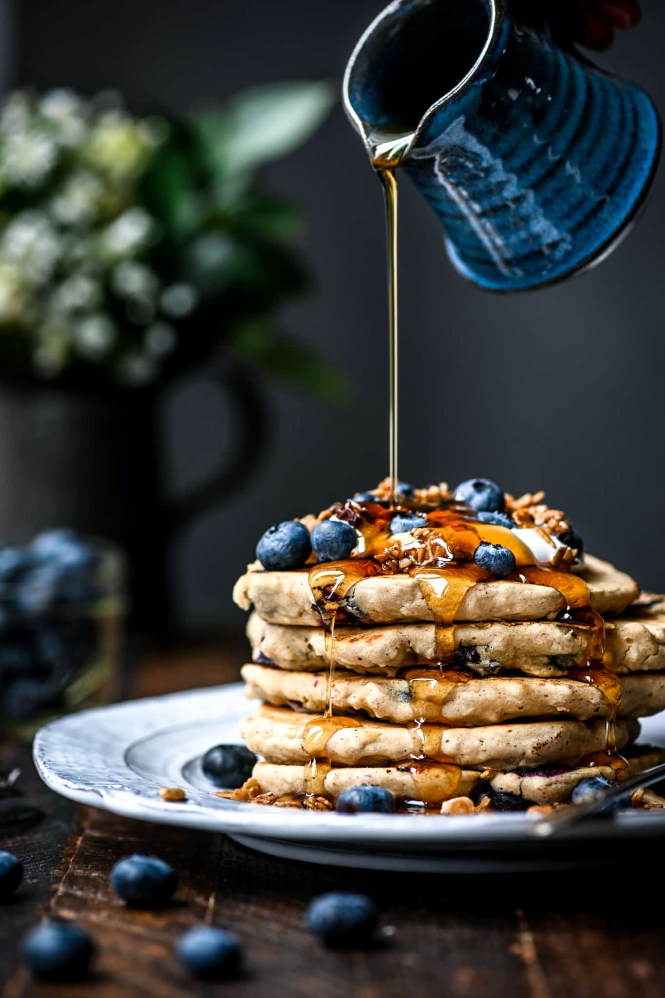 Side view of pouring maple syrup onto a stack of gluten free vegan blueberry pancakes with granola and fresh blueberries on a white plate