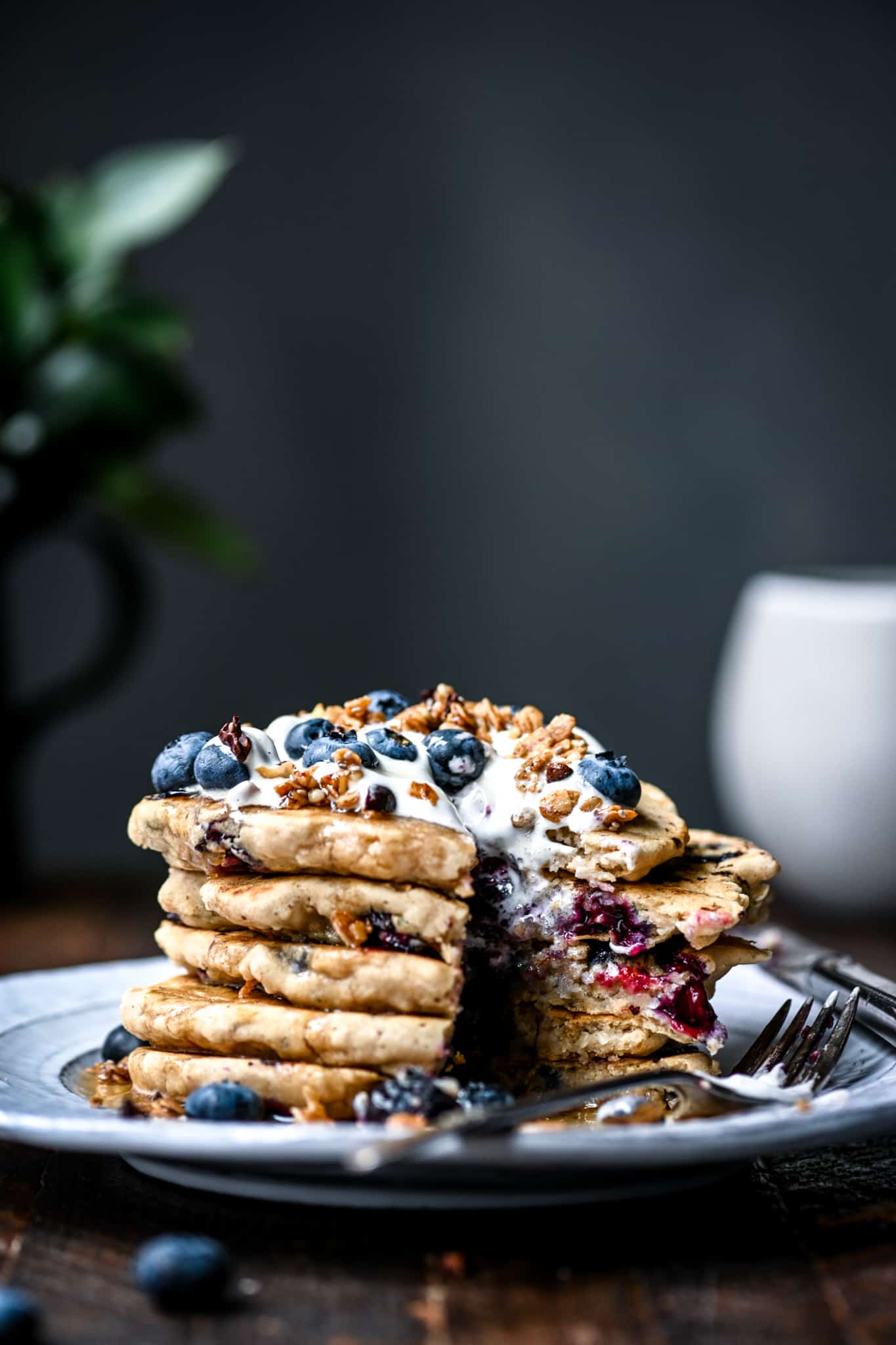 Side view of a stack of gluten free vegan blueberry pancakes with granola and fresh blueberries with a bite taken out on a white plate