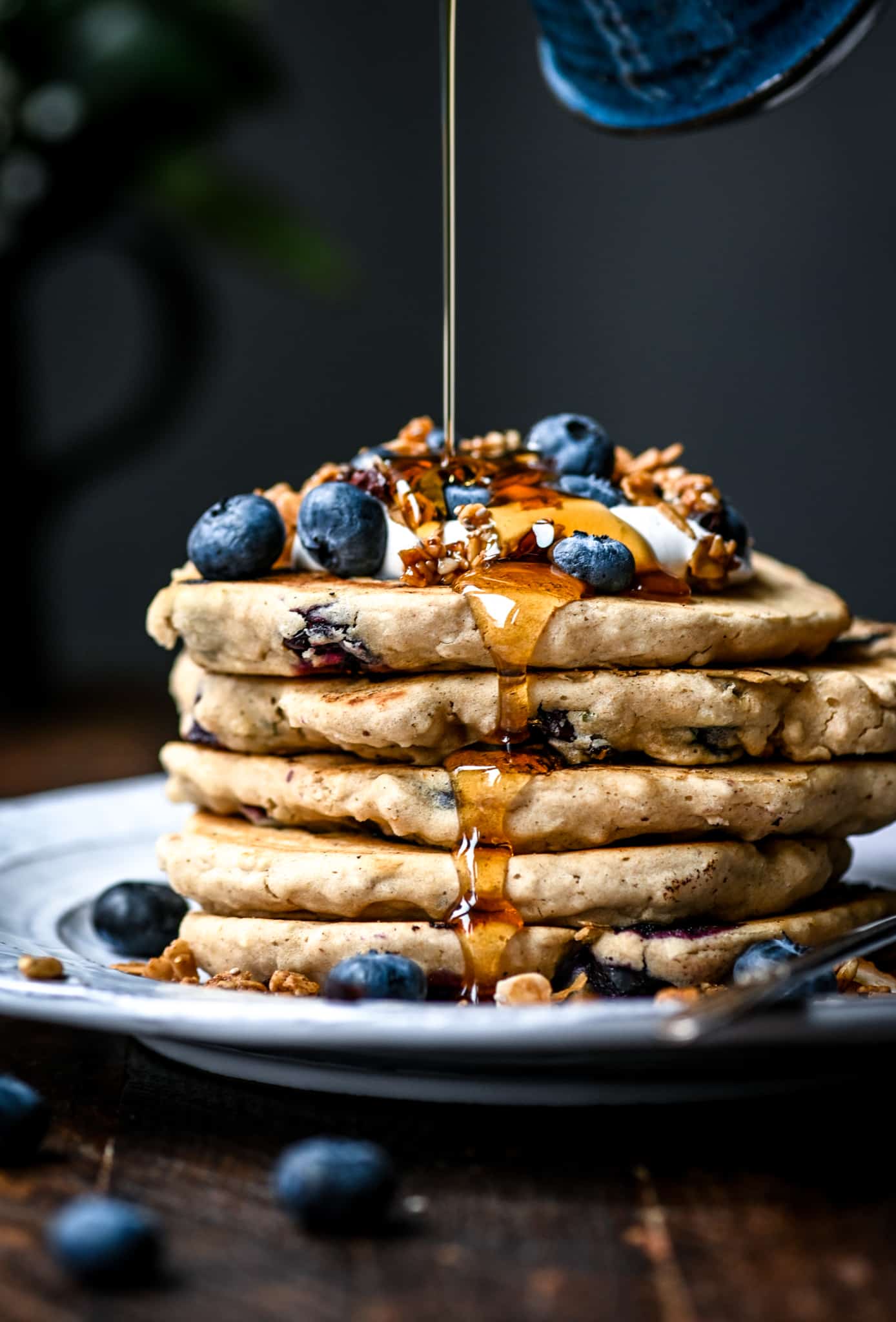 Side view of pouring maple syrup onto a stack of gluten free vegan blueberry pancakes with granola and fresh blueberries on a white plate for breakfast