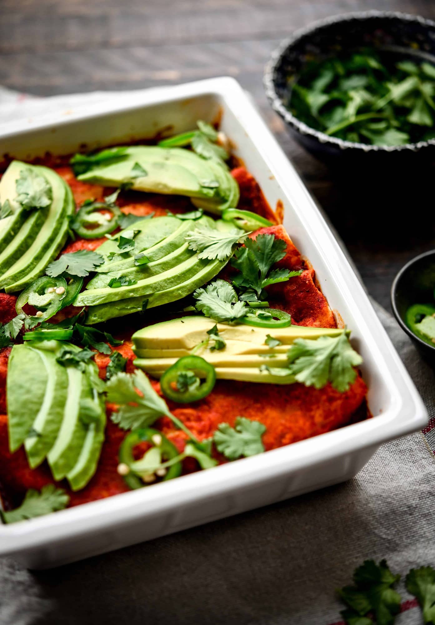 Side view of vegan enchiladas in white rectangular pan topped with avocado slices and mexican sauce