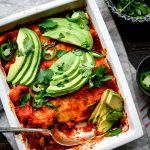 Overhead view of vegan enchiladas in white rectangular pan topped with avocado slices