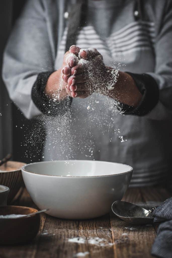 Person sifting gluten free flour from an antique sifter into white bowl on wood table