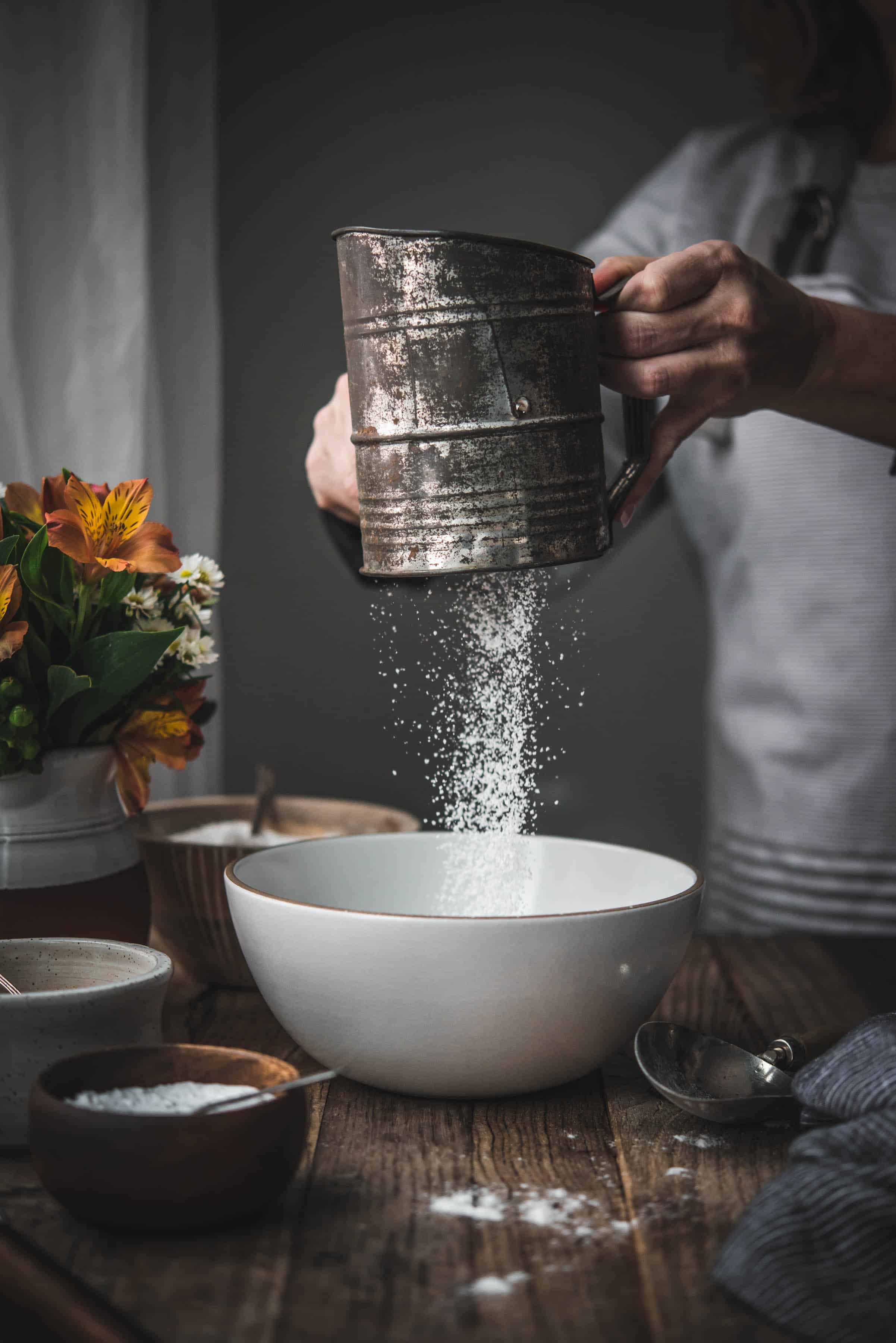 Person sifting gluten free flour from an antique sifter into white bowl on wood table